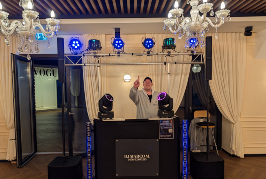 DJ Marco M hinter seinem DJ-Pult mit Lautsprechern, LED-Strahlern und beweglichen Lichteffekten bei einer Veranstaltung.