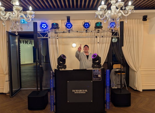 DJ Marco M hinter seinem DJ-Pult mit Lautsprechern, LED-Strahlern und beweglichen Lichteffekten bei einer Veranstaltung.