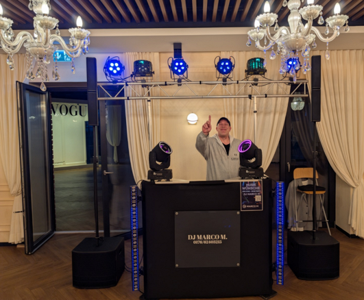 DJ Marco M hinter seinem DJ-Pult mit Lautsprechern, LED-Strahlern und beweglichen Lichteffekten bei einer Veranstaltung.