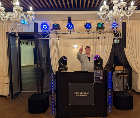 DJ Marco M hinter seinem DJ-Pult mit Lautsprechern, LED-Strahlern und beweglichen Lichteffekten bei einer Veranstaltung.