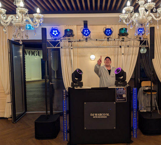 DJ Marco M hinter seinem DJ-Pult mit Lautsprechern, LED-Strahlern und beweglichen Lichteffekten bei einer Veranstaltung.