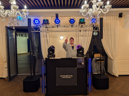 DJ Marco M hinter seinem DJ-Pult mit Lautsprechern, LED-Strahlern und beweglichen Lichteffekten bei einer Veranstaltung.